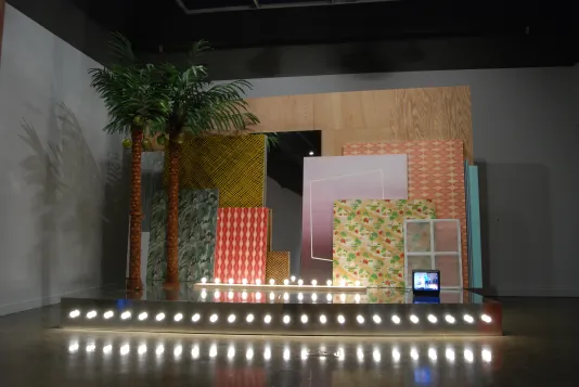 A setup with two fake palm trees, a red and white panel and other panels leaning on a plywood wall, a box monitor, row lights