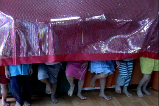 7 children whose faces and upper bodies are hidden under shiny red fabric stand in a row with legs and bare feet in mid-stomp.