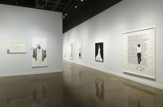 Installation view of the List Center gallery with work by Frances Stark hanging on the walls. In the foregound is the largest work, a block of text with the figure of an llustrated woman standing on a chair editing the words.