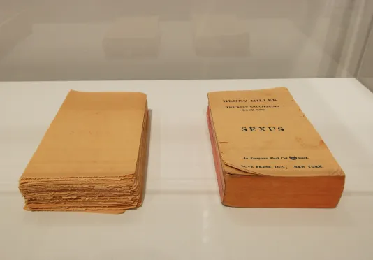 Close view of two books inside a glass vitrine. The left is an unbound copy with faded text. On the right is a bound copy with a cover that reads "Henry Miller, The Rosy Crucifixion Book One, Sexus." 