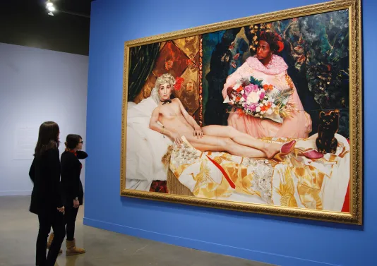 2 visitors view a large photo in a gold frame of a man portraying a nude woman lounging on a bed and a woman beside holding flowers. 