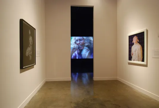 2 photos hang across from each other in a gallery that leads to a tall narrow entranceway where a video plays in a dark room.