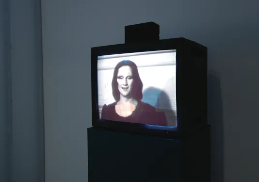 A video on a TV monitor shows a menacing looking figure with a painted white face, dark eyes, hair, lips, in a black dress.