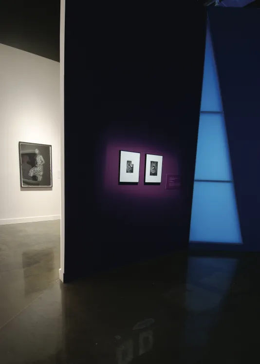 In a dark room 2 photos are illuminated against a dark purple wall, another artwork is hung in a well lit room to the left.