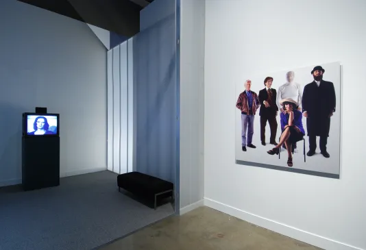 A monitor plays a video in a viewing room with plastic partitions, a photo of a group of people is mounted to the right.