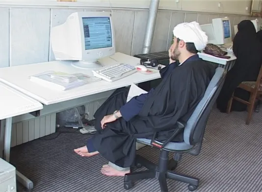 A barefoot man in a dark robe and white headscarf sits in an office chair at a workstation, looking at a computer screen.