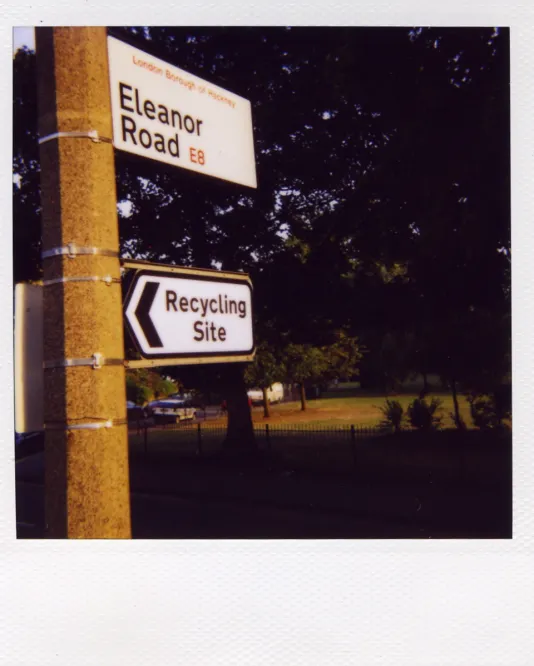 A white sign with “Eleanor Road” mounted above another sign “Recycling Site” on a concrete post before dark tree leaves