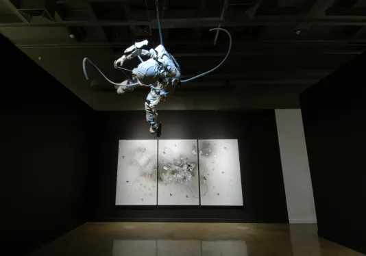 Cast resin and steel figure of astronaut dangles upside down above and before spot-lit triptych drawings on a black wall