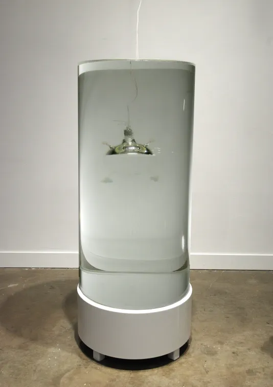 Nearly invisible glass rover tethered to white cord hovers in a large cylindrical tank filled with clear liquid.
