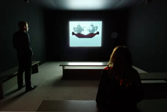 Two people stand in a dark gallery and view a projection of two white tea pots.