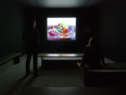 Two people stand in a dark gallery and view a projection of a closeup of pink food and lemon peel.