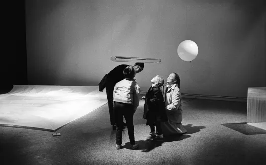 A photo of visitors viewing a clear cylindrical sculpture suspended in air from underneath it in Hans Haacke’s 1967 exhibition.