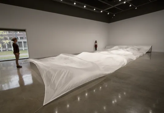 Visitors stand around a large sculpture made of a long white silk fabric installed over the floor that is wavy like the ocean. 