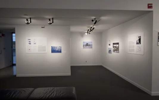 A small gallery of photographs and text panels mounted along several walls and lit from above with a bench in front.  