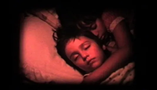 A still image of two children sleeping, heads on pillows, one’s arm around the other, the scene bathed in red light