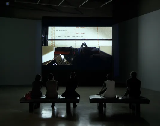 5 people sitting on benches in a dark gallery looking at a large movie screen with a still of a close up of a typewriter. 