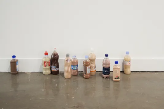 Ten plastic water or soda bottles on floor next to a wall, in various groupings and filled with beige and brown liquids