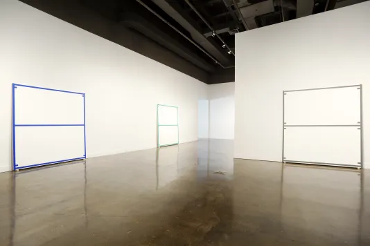 Three large canvases lean against two walls. Each canvas is blank except for being edged and bisected with a colored line.
