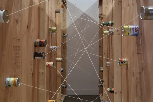 Food cans affixed on 2 facing walls, strings attached to each can connect to one another, criss crossing in the space between.