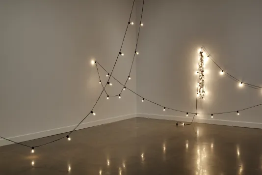 A lit string of bulbs scribbles across the image, reaching up out of frame then down, stretching and draping over two walls.