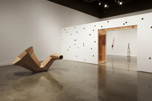 A gallery displaying a large cardboard funnel-like sculpture, a low perforated wall, and 2 objects on a wall in a room beyond.
