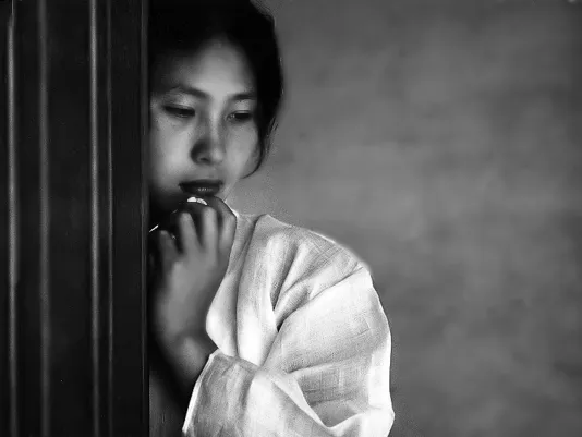 Muted mage of a young korean woman, in draped white clothing, cheek and hand resting against a wall, eyes cast down.