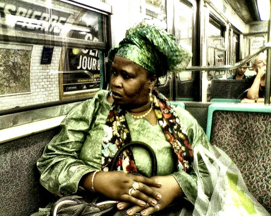 A dark skinned woman seated in a subway car, in jewelry, dressy clothing and headwear, holds a purse and stares out a window.