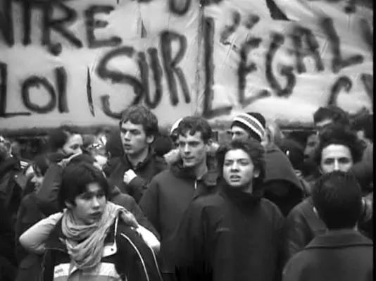 Black and white photo of several young people gathering in protest, a large handwritten sign filling the background.