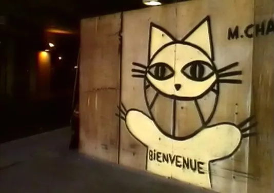 Comical stylized cat with a large grin and open arms, graffitied on a concrete wall. Bienvenue is written across its chest.