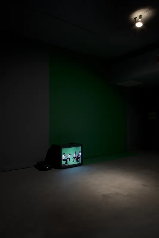 Displaying 4 musicians, a video monitor sits on a floor, spotlit by a ceiling lamp in a long shot of a dark room.