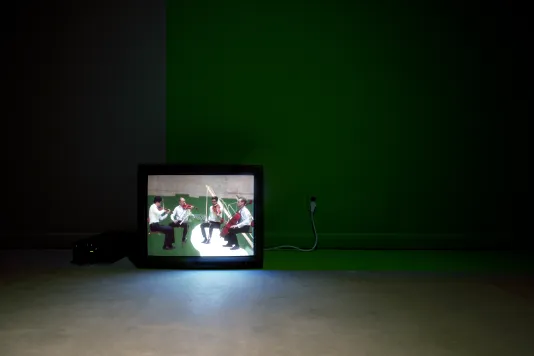 In a dark room, a video monitor sitting on the floor displays 4 seated musicians playing stringed instruments.