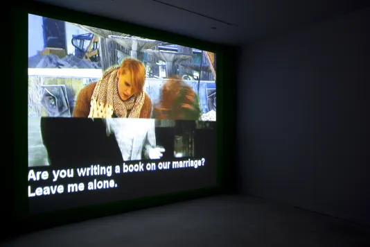 A video projection of a collaged image with white text running below, plays on a wall in an angled view of a dark room.