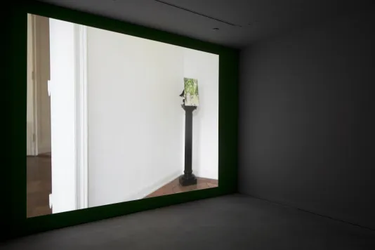 On a wall in a dark room, a video displays a pedestal holding an object tucked in the intersection of 2 white walls.