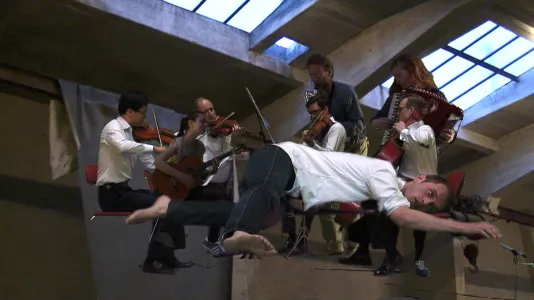 A man lies sprawled in front of a group of musicians, as they all hover in the air under a skylit architectural ceiling.