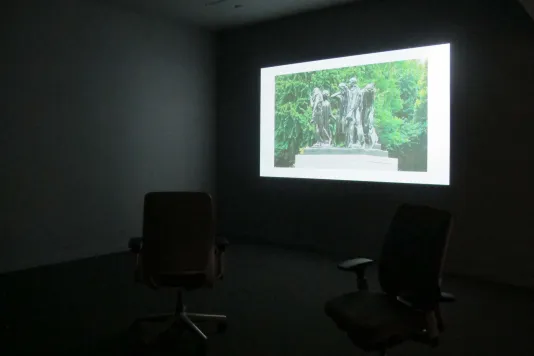 Two chairs hide in the darkness of the gallery with video installation lighting one wall