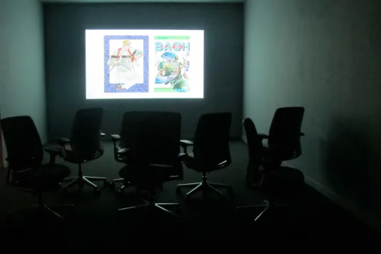 In a small, dark gallery space, 6 empty chairs sit in front of a brightly lit projection on the wall