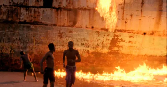 A wall of rust from an old ship creates a vivid scene as a line of flames rises near the base, silhouetting 3 young men.