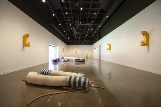 Long view of a space displays artwork including 5 aluminum hook shaped works on the walls, 2 fabric sculptures on the floor.