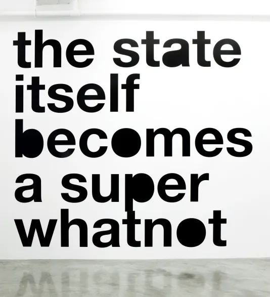 Large, black vinyl, lowercase Helvetica text, “the state itself becomes a super whatnot” on a white gallery wall.