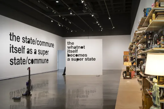 Large, black vinyl, lowercase text on 2 white walls, a vacuum cleaner in the foreground and on right, objects on wood shelves