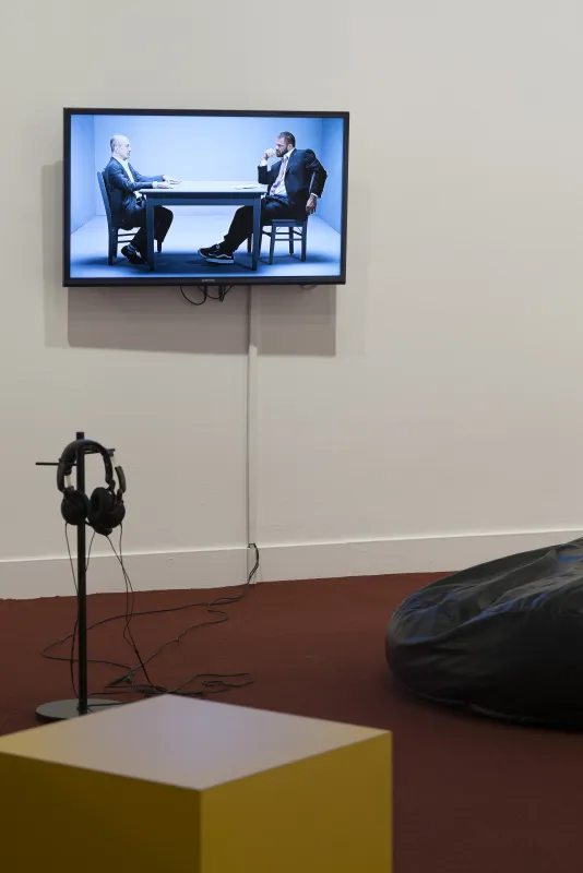 A wall-mounted monitor show 2 men sitting across the table from one another in a blue room, a headphone on a stand in front.
