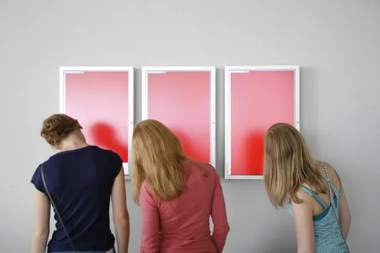 3 visitors with heads tilted to the left study the 3 red screens framed in silver and mounted on a white wall.