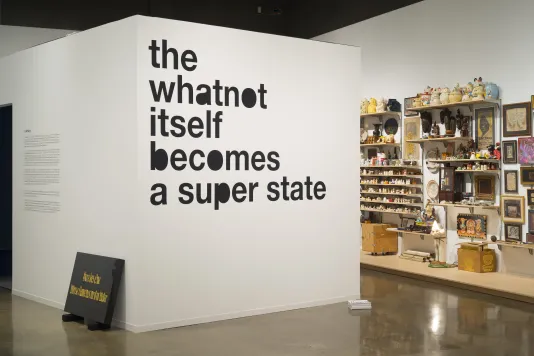 Black text “the whatnot itself becomes a super state”, a stone tablet propped against a wall and objects on shelves in back.