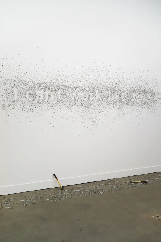 Nails on a white wall form the words “I can't work like this”, on the floor underneath, 2 hammers and scattered nails.