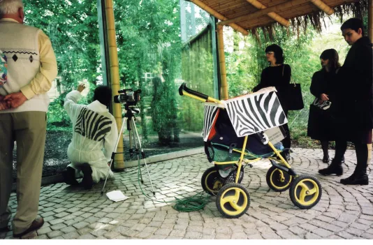 Zebra-pattern rectangles adorn the white overalls and cart of person kneeling next to a camera pointing out at green trees.