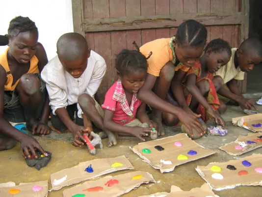 6 dark-skin children sitting, kneeling, squatting on the ground engage in a project with cardboard color paint palettes.