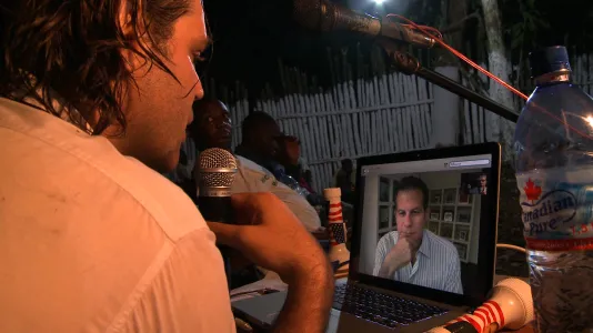 Lit at night outdoors, a man with shoulder-length hair in foreground speaks into a mic with a man on a laptop screen.