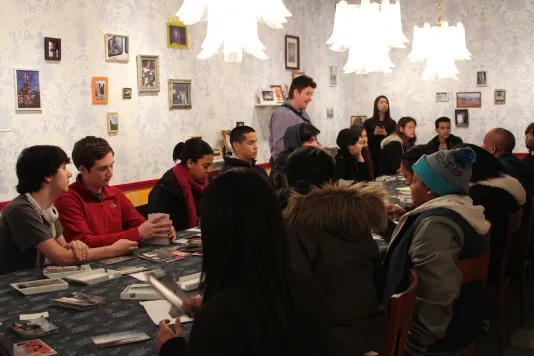 Students sit at a long table filled with photo trays, beneath 3 white hanging lamps. Framed photos dot patterned walls.