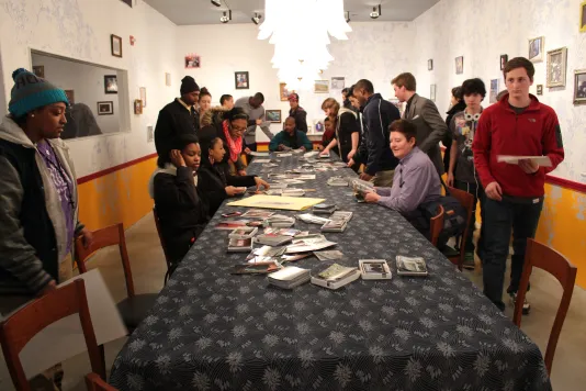 Long shot of a room where students mill about a cloth covered table filled with photos, petal shaped lamps hang, top center.