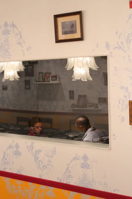 Beyond a window’s reflections, past a colorful patterned wall, a young man sits facing the artist in the adjoining room.